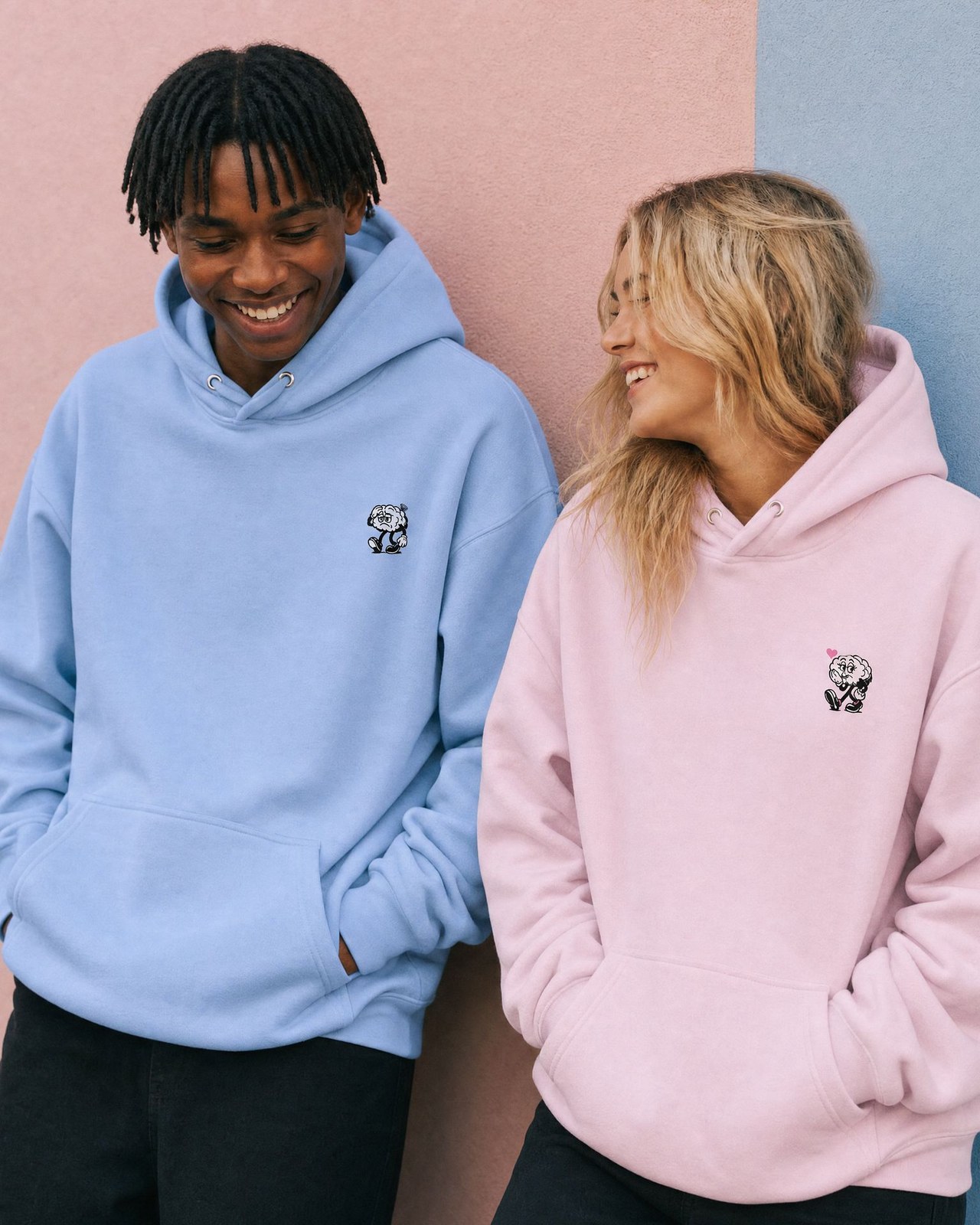 Two friends laughing in oversized hoodies against a pastel wall